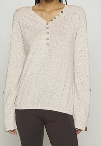 Light beige long-sleeve top with a V-neck, featuring a front button placket and brown dot pattern. Elastic hem and relaxed fit.