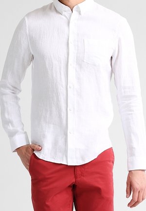 Homme portant une chemise blanche à manches longues boutonnée et un pantalon rouge, une main dans la poche et l'autre détendue sur le côté.