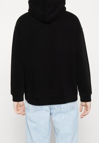 Sweat à capuche noir avec une poche kangourou, des poignets et un ourlet côtelés. Vue de dos montrant une coupe décontractée et une texture douce. Associé à un jean bleu clair.