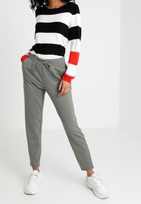 Zwart-wit gestreepte lange mouwen top met rode accenten, gecombineerd met grijze taperende broek en witte sneakers. Gladde textuur, casual pasvorm.