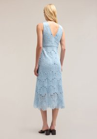 Rochie din dantelă de culoare albastru deschis, cu spate în V, modele tăiate intricate și un tiv de lungime medie. Asortată cu sandale cu toc bloc negre.