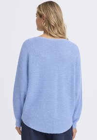 Femme aux cheveux blonds portant des boucles d'oreilles argentées en forme d'anneau et un pull en tricot bleu clair, vue de dos sur un fond uni.