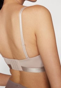 Lys beige bralette med tynde stropper, en glat stoftekstur og et satinlignende accentbånd. Har justerbar hægte- og øjentilslutning på bagsiden.