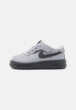 Baskets basses blanches et grises Nike avec logo Swoosh noir, lacets et bride Velcro, semelle extérieure en caoutchouc noir.