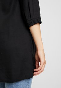 Musta tunika, jossa on käännetyt hihat, ja se on valmistettu sileästä kankaasta, jossa on kaareva helma. Kangas vaikuttaa kevyeltä ja pehmeältä.