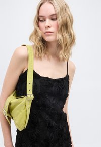 Νεαρή γυναίκα με κυματιστά ξανθά μαλλιά που φοράει μαύρο φόρεμα με υφή λουλουδιών και κρατάει μια υφασμάτινη τσάντα ώμου σε ανοιχτό πράσινο χρώμα.