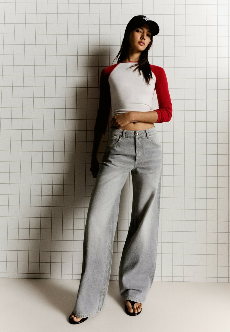 Jeune femme portant un crop top rouge et blanc à manches longues, un jean gris à jambes larges, des tongs noires et une casquette de baseball noire, debout contre un mur carrelé.