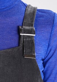 Svart kordfløyelsoverallrem med metallspenne bært over en blå ribbet langermet skjorte på en persons skulder og bryst.