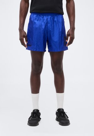 Homme portant un short de sport bleu brillant, des chaussettes blanches et des chaussures de course noires, debout sur un fond uni.