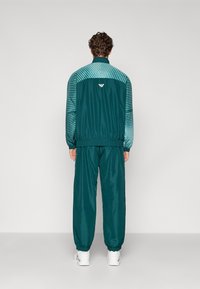 Tiffany-kék tréningruha csíkkal átszínezett dizájnnal a dzseki vállán, rugalmas mandzsettákkal és derékpánttal, amelyhez illeszkedő, tapered nadrág tartozik.