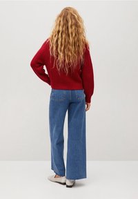 Czerwony, dziany sweter, szerokie niebieskie dżinsy z postrzępionym dołem i jasne buty. Włosy są długie i faliste, na jednolitym tle.