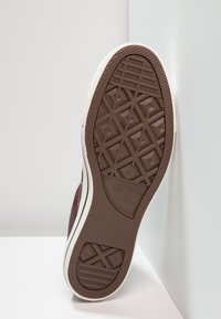 La semelle de sneaker d'un marron foncé présente un motif de tread géométrique avec des surfaces texturées. Un bord en caoutchouc blanc entoure la semelle, étiquetée "Converse All Star."