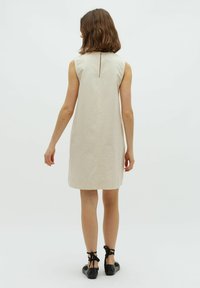 Beiges ärmelloses Kleid mit einer Rückenschlitzdetail, knielang, aus einem strukturierten Stoff, kombiniert mit schwarzen Riemchenschuhen ohne Absatz.