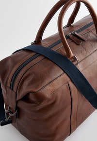 Brun lædertaske med en tekstureret overflade, robuste håndtag og en marineblå skulderrem. Har en lynlås lukning langs toppen.