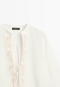 Blouse blanche avec encolure ronde, broderie florale beige le long de l'ouverture devant et fermetures à nouer blanches sur la poitrine.