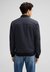 Marineblaue Bomberjacke mit geripptem Kragen und Bündchen, die eine glatte Textur und ein einfaches Design aufweist. Kombiniert mit hellblauen Jeans.