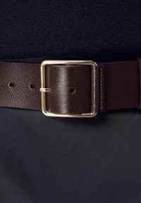 Cintura in pelle marrone con superficie texturizzata, dotata di una fibbia in metallo dorato e cinque fori per la regolazione. Sfondo in tessuto nero.
