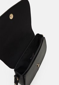 Borsa a mano in pelle nera con superficie texture, accessori in oro e un interno spazioso. Chiusura a pattina con bottone a scatto. Gli accenti sono minimi.