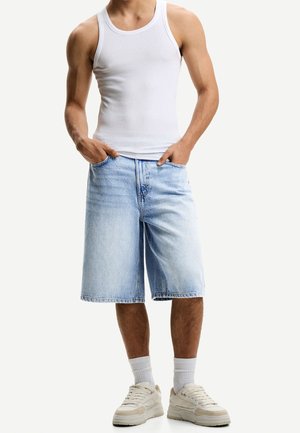 Homme portant un débardeur blanc, un short en jean bleu clair, des chaussettes blanches et des baskets blanches, debout les mains dans les poches.