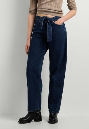 Personne portant un jean large taille haute bleu foncé avec une ceinture en tissu, un crop top marron côtelé, et des bottines noires avec détail chaîne.
