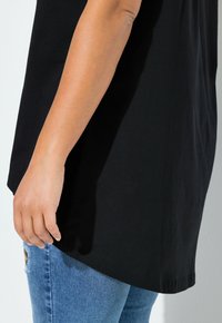 Bras et main d'une personne portant une chemise noire ample et un jean bleu avec un léger détail usé sur la cuisse.