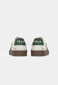 Sapatilhas brancas com detalhes verdes, feitas de couro, com uma textura suave, bico arredondado e sola de borracha castanha. Logótipo "VEJA" em dourado.