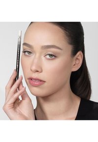Zwarte wenkbrauwpotlood met een witte punt, vastgehouden door een persoon met een lichte huid, steil donker haar en subtiele make-up, tegen een neutrale achtergrond.