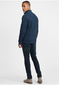 Tengeréskék bordázott pulóver magas gallérral, sötét farmernadrággal és világosszürke csizmával párosítva. Az anyag texturált mintázatot mutat.