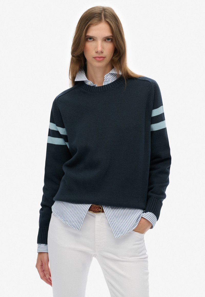Marineblauwe trui met lichtblauwe strepen op de mouwen, ronde halslijn, gedragen over een lichtblauw en wit gestreept shirt, gecombineerd met witte broek.