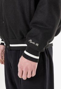 Sort uldblandet varsity-jakke med hvide stribede manchetter og talje. Har broderet logo på manchetterne. Nærbillede af armen.