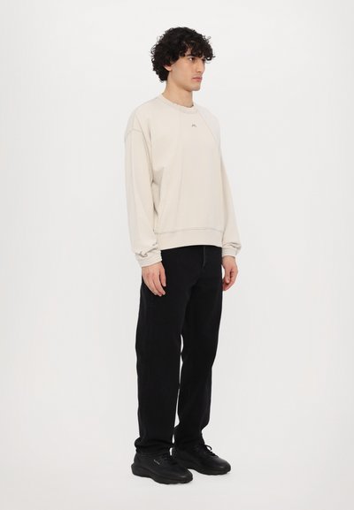 Sweatshirt beige clair avec des manches raglan, des poignets côtelés et un logo sur le devant, associé à un pantalon noir ample et des baskets noires.