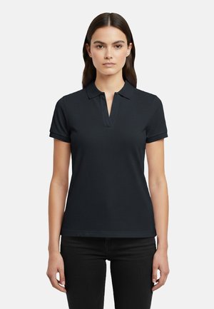 Donna in piedi di fronte, che indossa una polo nera a maniche corte e pantaloni neri su uno sfondo bianco uniforme.