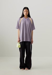 Chemise oversized lavande avec des accents beiges et un décolleté en V ; assortie à un pantalon large noir et à un petit sac à main vert clair.