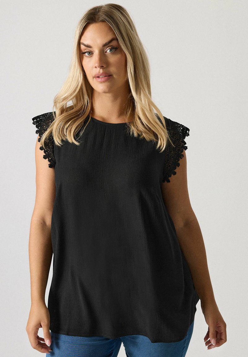 Blonde Frau trägt ein schwarzes ärmelloses Top mit Spitzenärmeln und blaue Jeans, steht vor einem hellen Hintergrund.