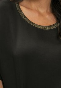 Haut noir avec un col rond présentant une bordure dorée scintillante. Le matériau semble lisse et léger. Les manches courtes sont visibles.