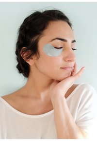 Hydrogel-Augenmaske in Hellblau unter dem linken Auge einer dunkelhaarigen Frau, die ein weißes Oberteil trägt, vor einem hellen Hintergrund.