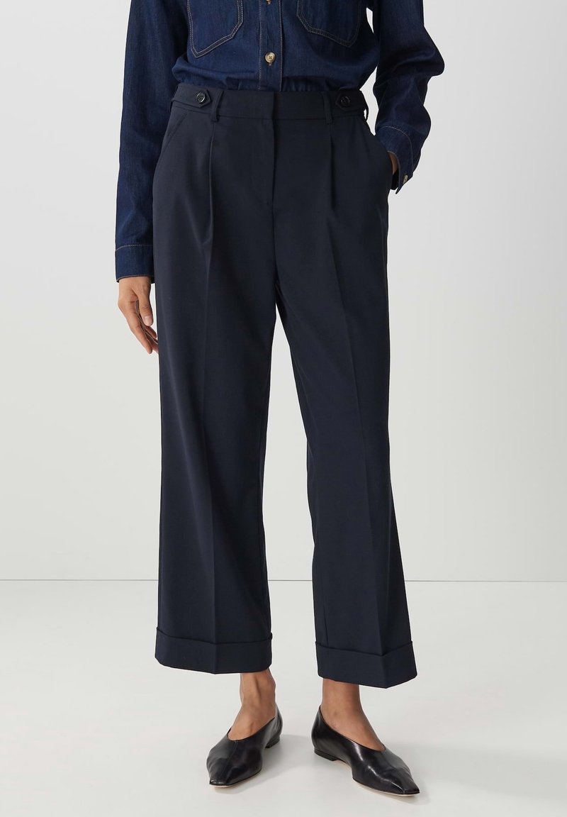 Marina höga byxor med veck, tillverkade av slätt tyg, med sidsfickor och uppvikt fåll, parat med svarta slip-on skor.