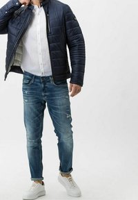 Chaqueta acolchada navy con cuello alto, camisa blanca de botones, jeans azules desgastados y zapatillas blancas, mostrando un ajuste relajado.