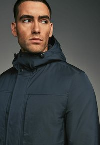 Chaqueta impermeable azul marino con un alto cuello, capucha ajustable, bolsillos frontales y costuras en contraste. Textura de tejido suave y funcional.
