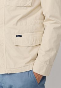 Beige jas met mouw en zak met label, hand in lichtblauwe broekzak, focus op structuur van de stof en stikdetails.