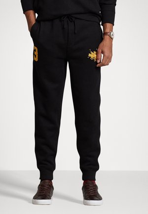 Hombre con pantalones deportivos negros con el número dorado "3" en el muslo izquierdo y el logo dorado de jugador de polo en el muslo derecho, combinado con zapatillas oscuras.