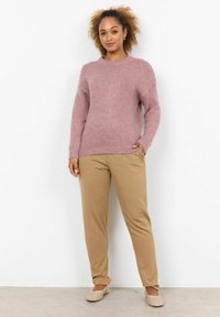 Light pink knitted sweater with round neckline. Beige tapered pants with a smooth texture. Model wearing textured slip-on shoes.