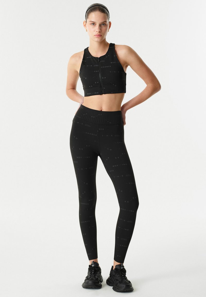 Černý sportovní crop top se zipem a odpovídající legíny s vysokým pasem, obojí s opakujícím se textovým vzorem. Měkký, elastický materiál.