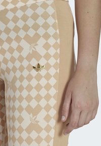 Beige legíny s diamantovým vzorem, zdobené zlatým logem Adidas. Měkký materiál s elastickým střihem a kontrastními světlejšími bočními panely.