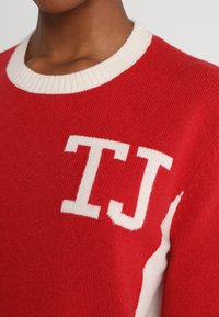 Osoba nosząca czerwony sweter z dzianiny z białym wykończeniem i białymi literami "TJ" wydzierganymi na piersi.