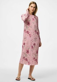 Rosa plisserad klänning med blommönster i lila och grönt. Långa ärmar och rund halsringning, når till mitt på vaden. Bärs med svarta flip-flop-sandaler.