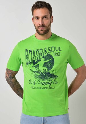 Mężczyzna z wytatuowaną ręką, ubrany w jasnozieloną koszulkę z ciemnoniebieską grafiką surfingową i napisem „Board & Soul Surf Supply Co., Echo Beach, Bali.”