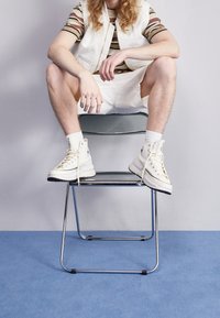 Hochschnittige Sneaker aus cremefarbenem Canvas mit weißen Schnürsenkeln und schwarzem Gummisohle. Modell sitzt auf einem transparenten Stuhl, präsentiert auf einem blauen Teppich.