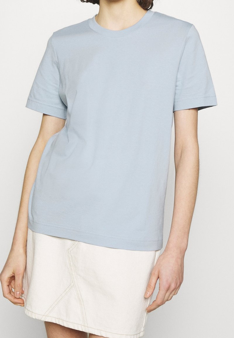 Camiseta de algodón azul claro con mangas cortas, cuello redondo, corte relajado, tela ligeramente texturizada, llevada sobre una falda de mezclilla blanca.