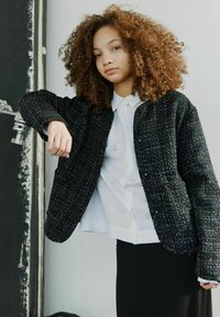 Črna teksturirana jakna z tkanim vzorcem, obleka nad belo srajco z diskretnim vezenjem, v kombinaciji s črno krilom. Preprost, sproščen dizajn.